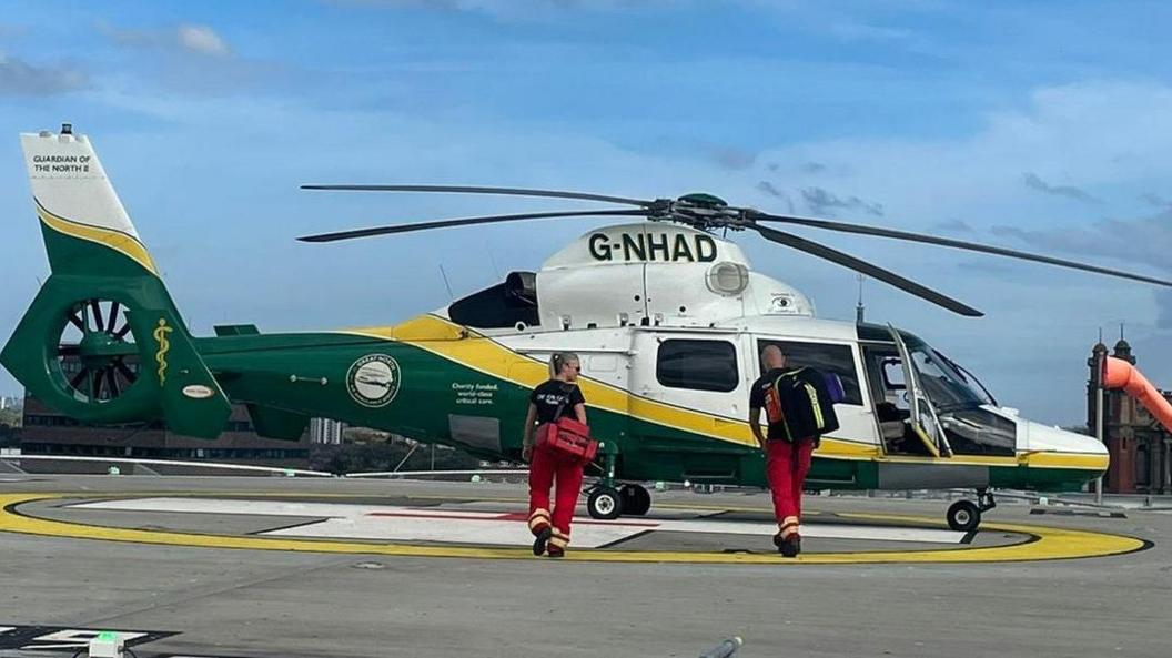 A GNAAS helicopter parked on a helipad with two paramedics walking towards it, carrying medical bags. The skyline of a city is visible behind the helicopter, set against a blue sky with a few white clouds. The helicopter is white, yellow and green. The paramedics are wearing dark t-shirts and red trousers. The helipad is a concrete platform on a rooftop with a yellow and white symbol painted on it where the helicopter is parked.