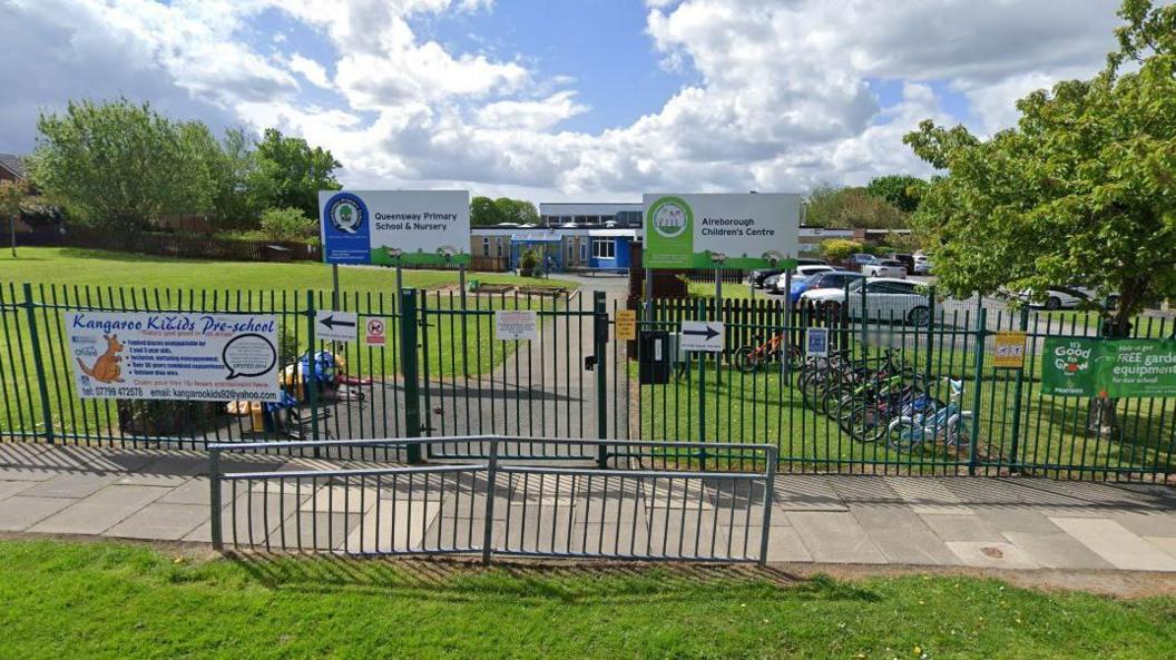 The outside of a primary school showing railings and a gate, some grass in front and several small bikes lined up in the playground 