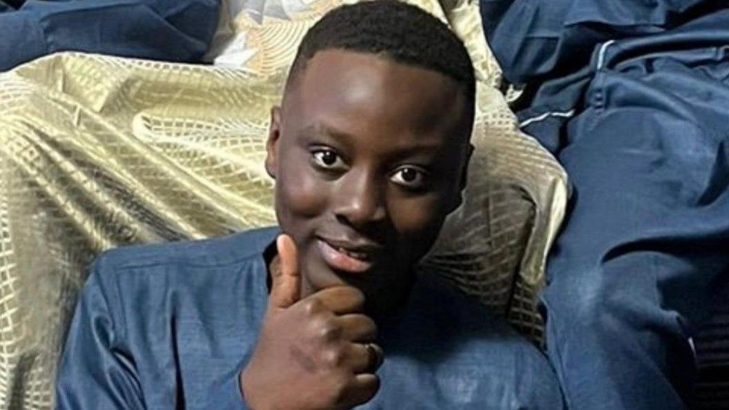 A young teenager sits smiling and giving a thumbs up to the camera.