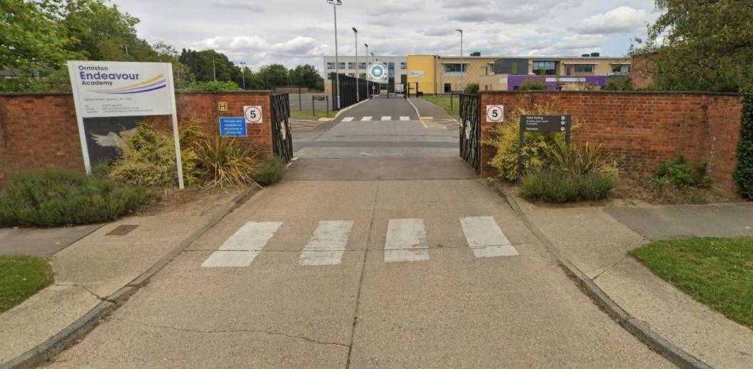 Front of Ormiston Endeavour Academy with a sign on left side of a wall with a road going through a gate. 