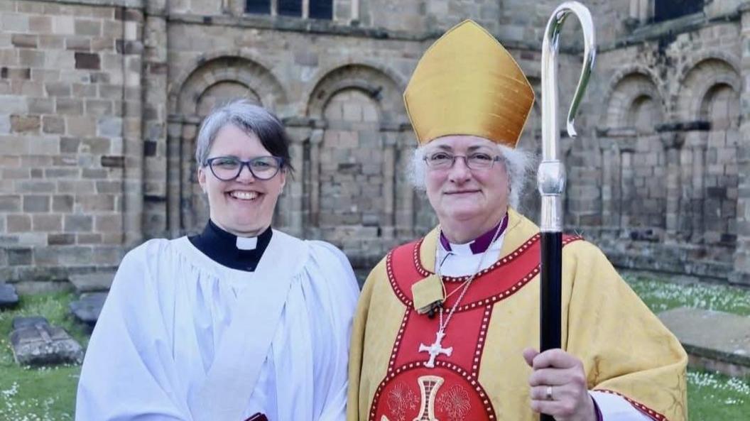 On the left is Donna Robinson wearing a white dalmatic and stole, with short grey hair, and glasses, smiles at the camera. She also has a dog collar on. To her right, is Sarah Clark, with glasses and grey hair, wearing a Bishop mustard hat and dalmatic with a cross, holding a bishop staff. They are standing in front of a stone church.