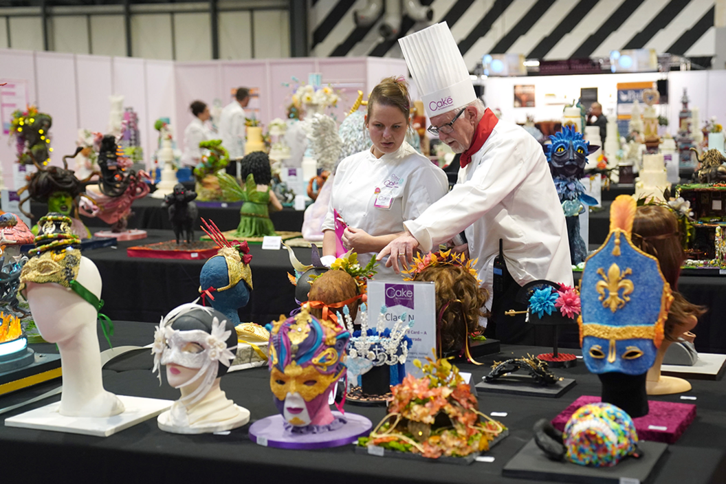 Two judges in white chef's outfits confer as they inspect several of the cakes depicting heads wearing different masks