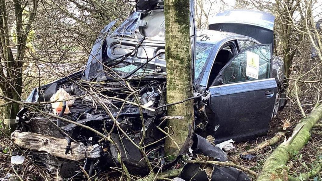 The front of a car smashed up with its bonnet off and in the air. Its passenger front door and boot is open. A tree is stood against the front left of the car.