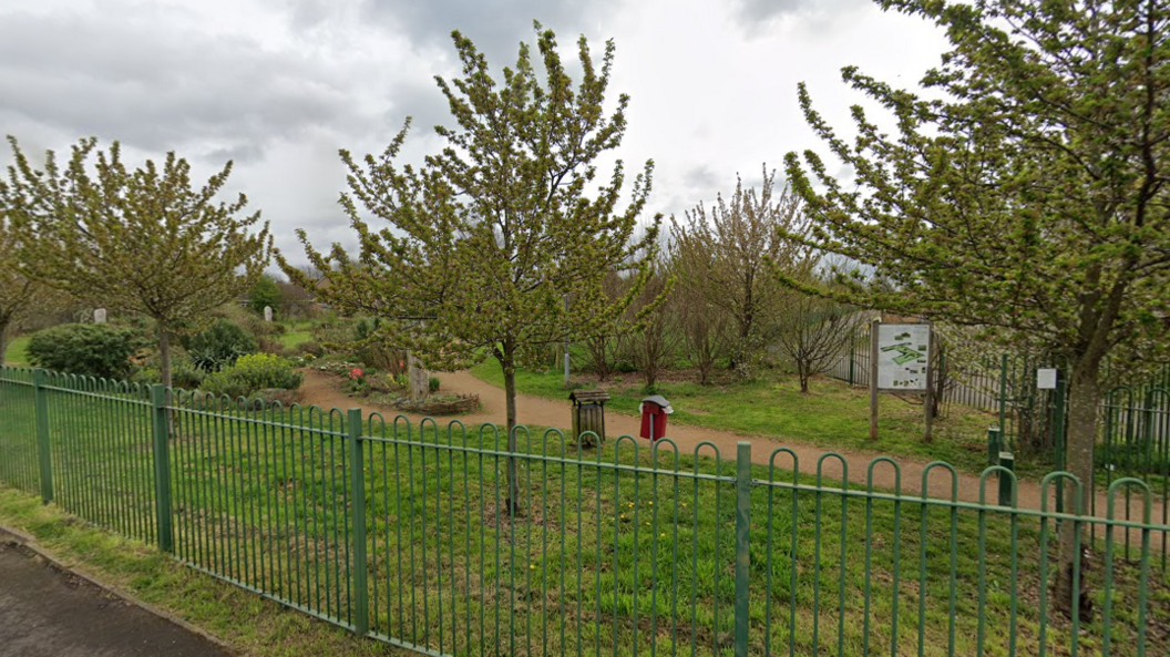 A street view image of a small grass park with small trees and a brown footpath running through it. The photo is taken from outside the park. There are metal railings on the edge of the park