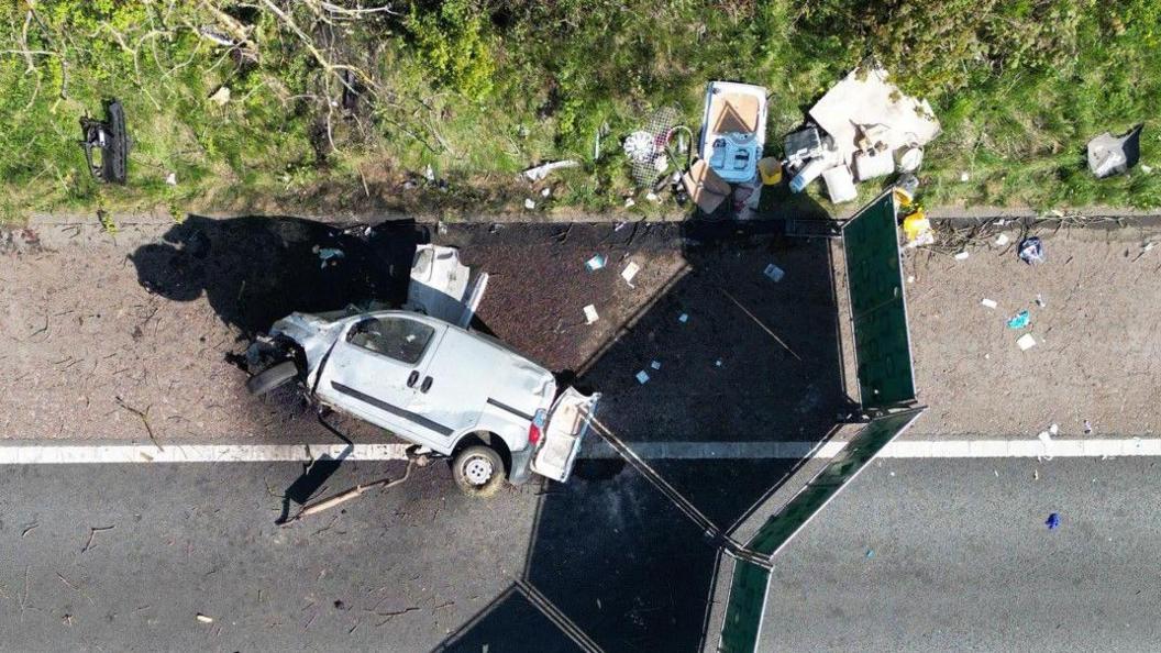 An aerial drone shot of the van, lying on its side on the hard shoulder of the motorway. Its bonnet is badly damaged and the passenger side front wheel has been torn off. There is other debris littering the grass verge, and a screen has been put up around the scene.