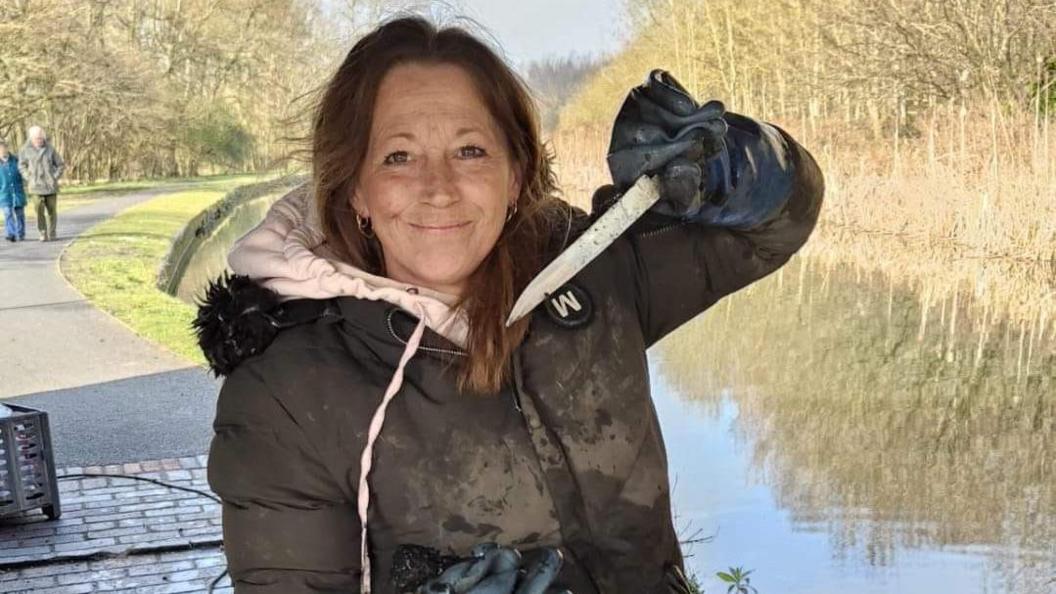 Gill Shilvock holding a knife that she has pulled out of a canal, with the waterway in the background