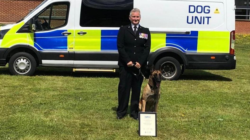 A police dog handler and a Belgian Malinois dog standing in front of a police van with a framed certificate placed in front of them