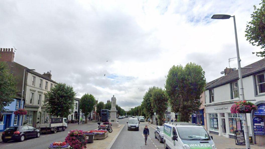 Cockermouth's Main Street with cars and vans travel on either side of the road. There are a number of shops along the street and a statue in the distance.