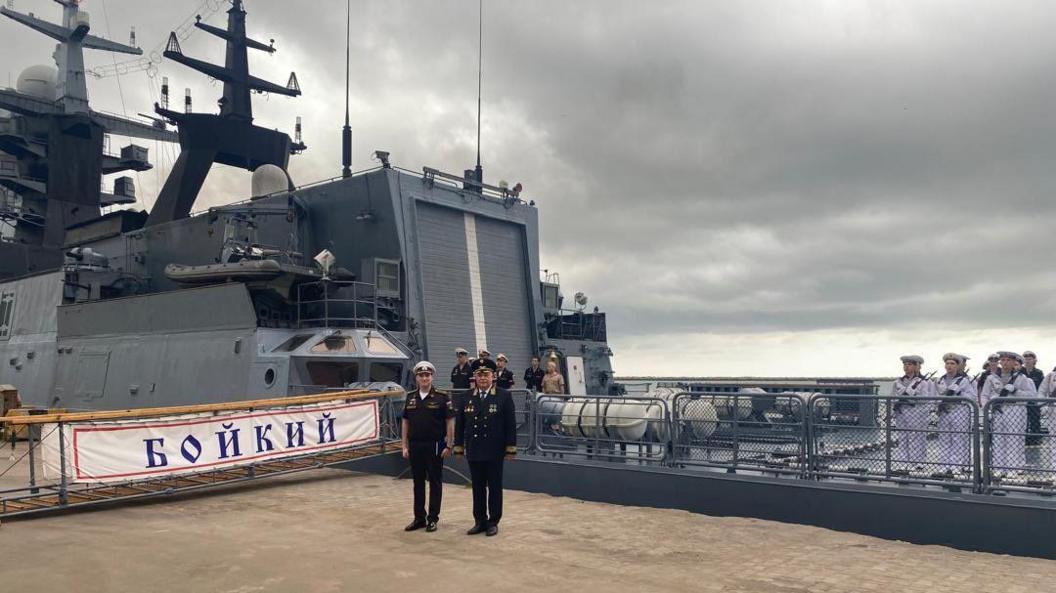 Russian naval officer standing on the deck of the Boikiy. A banner with the ships name can be seen in the background. Two officers are seen wearing uniforms, while enlisted men carry rifles.