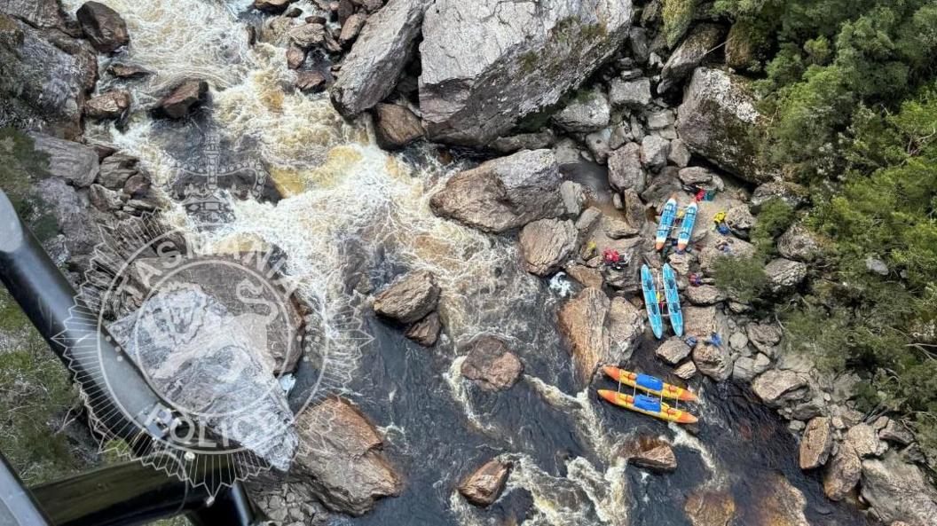 Aerial photo shows part of the Franklin River in Tasmania. There are rocks in the river and six blue and orange kayaks are placed on the rocks. The river looks rapid and is surrounded by vegetation