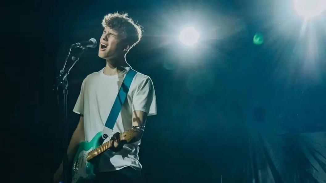 Ben from Revivalry playing his guitar and singing on stage. The guitar is mint green and he is wearing a white t-shirt and is surrounded by bright lights