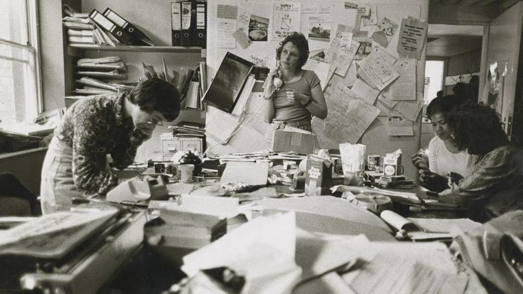 A black and white image of four women sitting around a desk. Two are on the phone and two are lighting cigarettes.