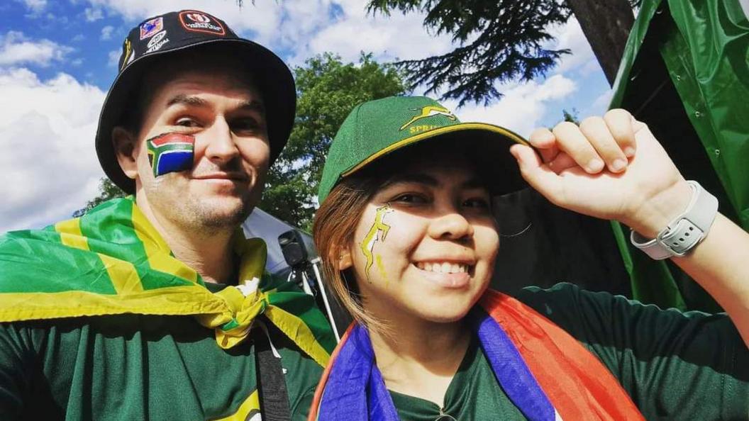 A man and a woman wearing South African rugby shirts, the man with a black bucket hat on and the South African flag painted on his cheek, a South African flag tied around his neck, with the woman wearing a green rugby hat, a Springbok painted on her cheek with a Philippines flag around her neck, posing outside on a cloudy day,