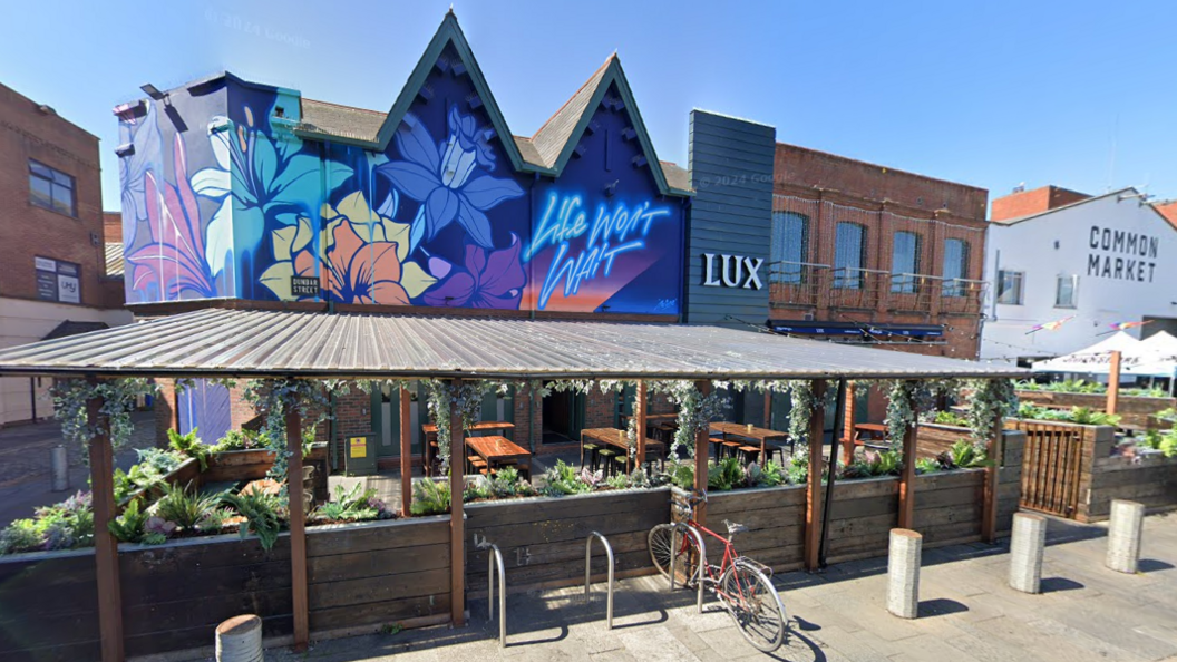 The exterior of Lux nightclub in Belfast. It shows a colourful mural wall with 'life won't wait' in neon-style spray paint. There is also a sheltered beer garden.