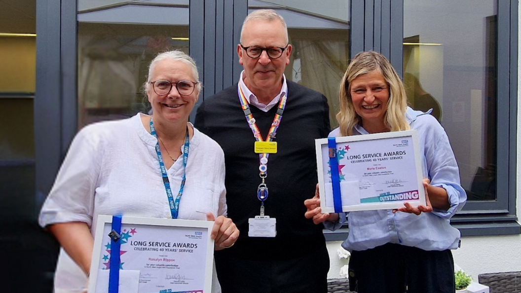 Marie and Ros holding their framed award- standing on either side of Steve Barnett- who is wearing a black jumper and a pink collar shirt.