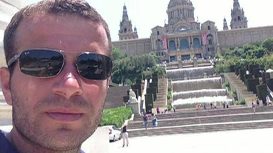 Albert Xhediku poses for a selfie in front of the Palau Nacional in Barcelona, a palace with domed turrets behind a cascading water feature and steep steps down to where he is standing. Mr Xhediku has short, brown hair and a close-cropped beard and is wearing sunglasses.