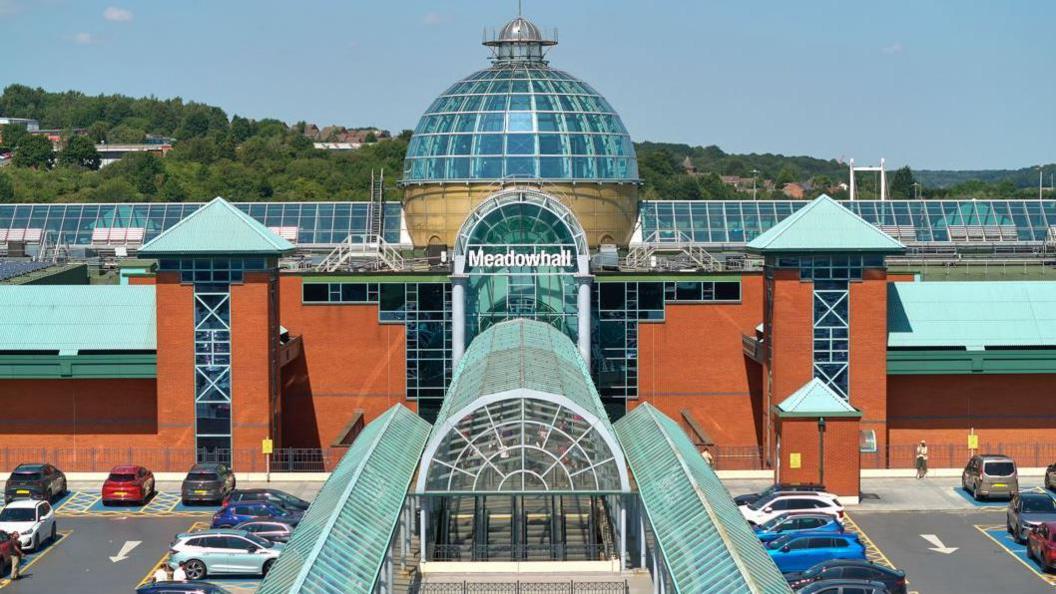 An orange fronted building with glass detail including a large glass dome. There is a sign in white writing which says 'Meadowhall' and surrounding the building is a car park with parked cars.