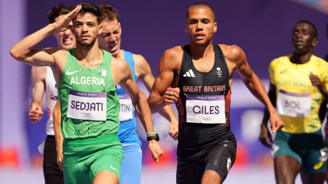 Five male athletes running. On the left a man in a green Alergia uniform salutes, to his right is Elliot Giles, wearing a black vest that says Great Britain