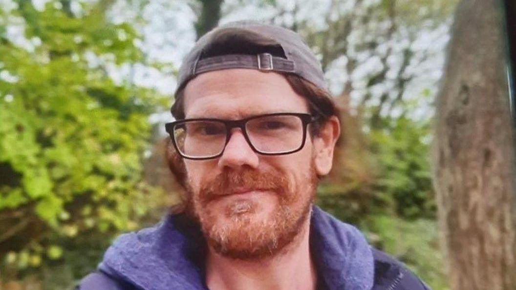 Shane Lynch pictured wearing a navy blue hoodie and navy blue jacket. He is also wearing a grey cap and has black square glasses. He also has long ginger hair and a ginger beard.