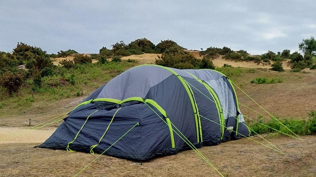 A large grey tend with bright green stripes and guide roles is pitched in the New Forest. It is surrounded by dry looking grass and vegetation.
