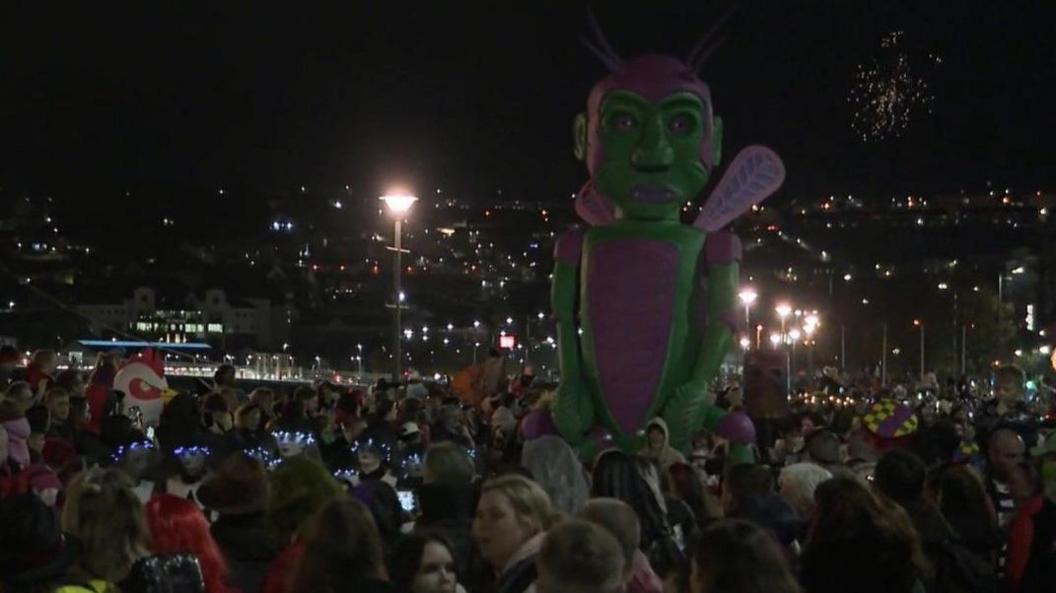 A large green and purple fly-like creature flanked by huge crowds either side of the parade 