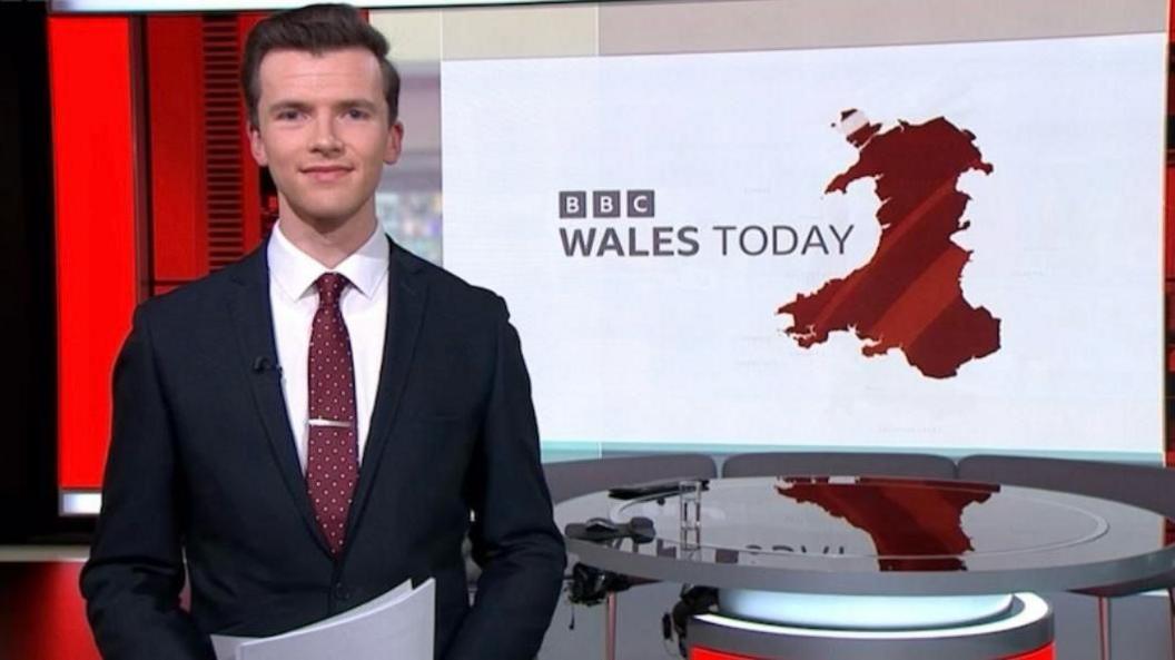 Daniel Heard wearing a suit, shirt and tie and standing in the BBC Wales Today news studio with the desk and screen behind him