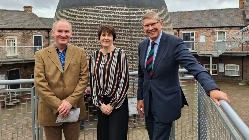 A woman is standing outdoors in between two men. They are positioned against a set of metal railings and looking towards the camera. Behind the group is a bottle-shaped bottle kiln made of brick and a series of two-storey factory buildings.