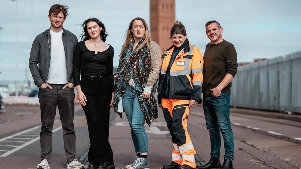 A group of five men and women, four in casual clothing and one in hi-vis safety wear, pose for a publicity photo in front of Grimsby's dock tower.