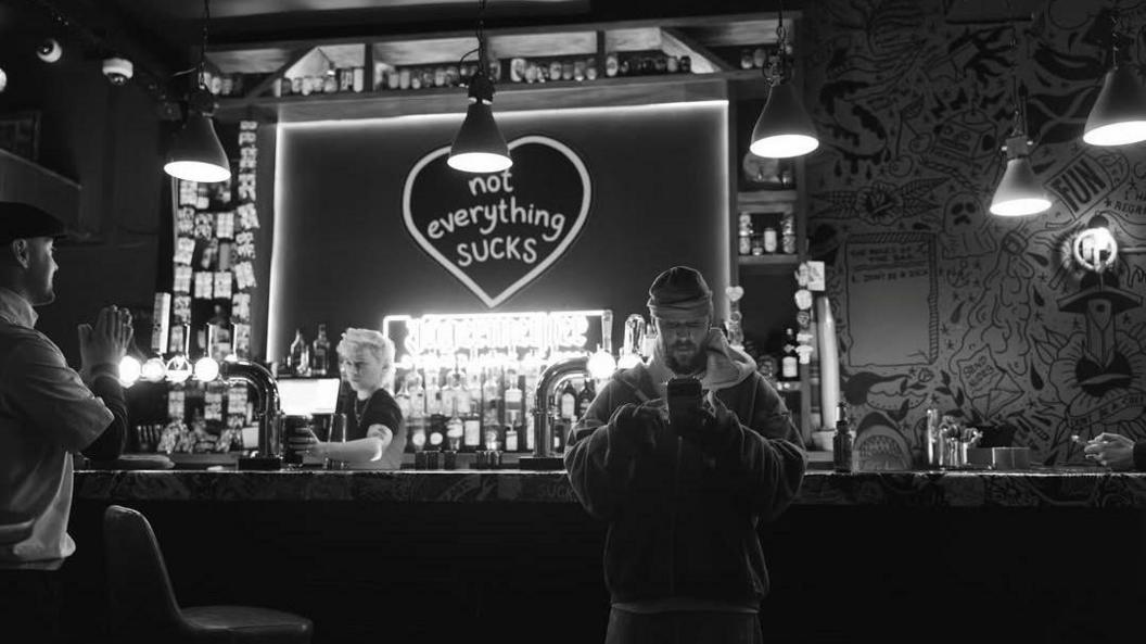Justin Bieber leaning against a bar, looking at his phone. A graphic behind the bar says "not everything sucks" inside a heart. A barman is serving a pint to another customer. The image is in black and white. 