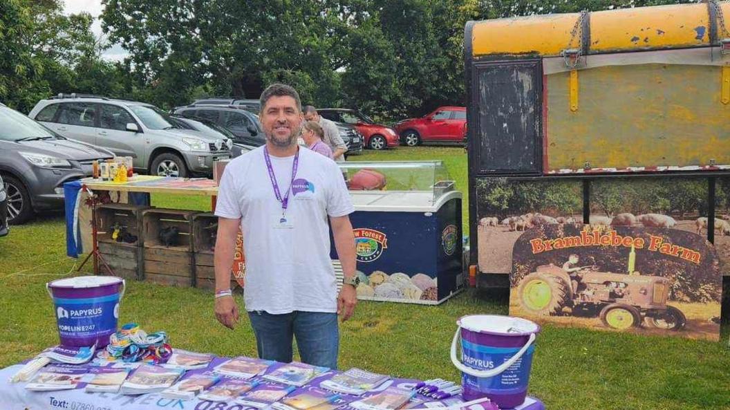 A smiling Trevor behind a stand with Papyrus leaflets on it, in a field with signs that read "Bramblebee Farm"