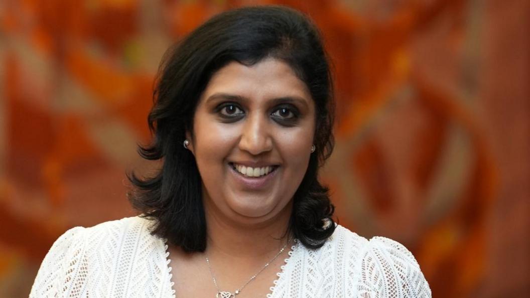 Priyanka Pandya-Bhatt, wearing a white outfit, looking at the camera and smiling, she has shoulder length dark hair, and is wearing a silver necklace. She is front of a red wall. 