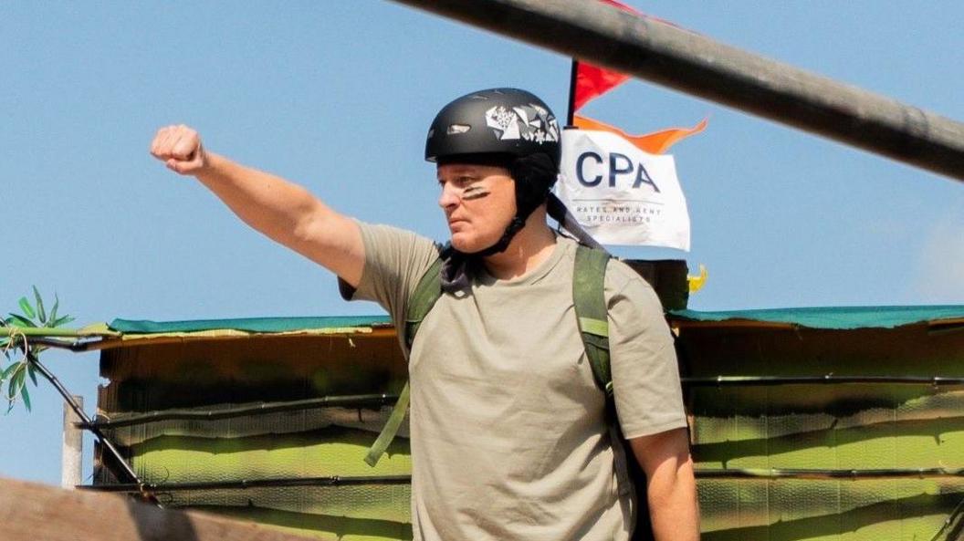 A man wearing a beige T-shirt and a black crash helmet salutes the crowd as he gets ready to take part in the Birdman Bristol event