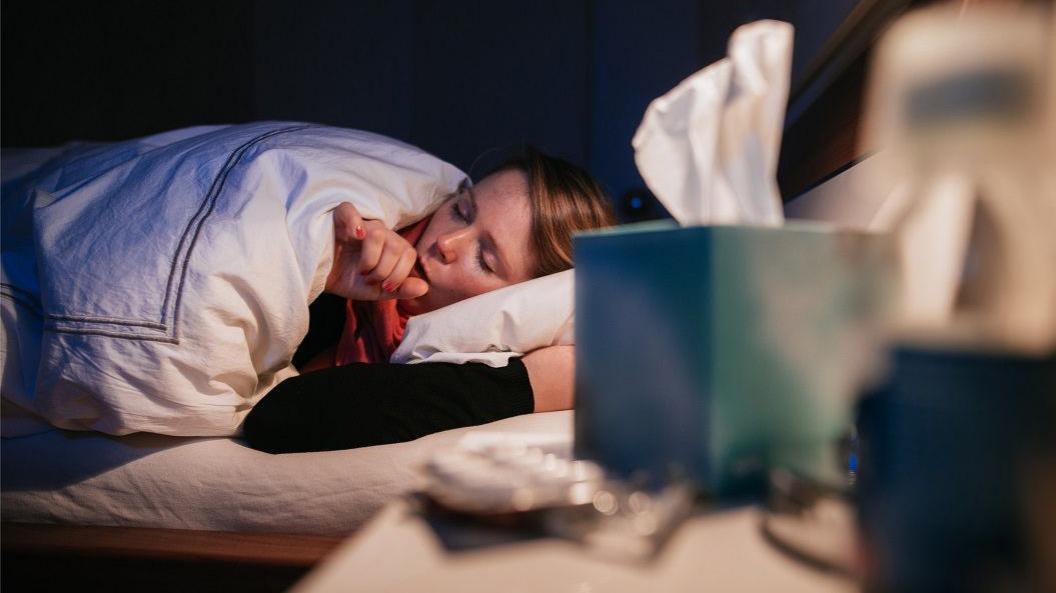 Woman in bed coughing. There are tissues nearby.