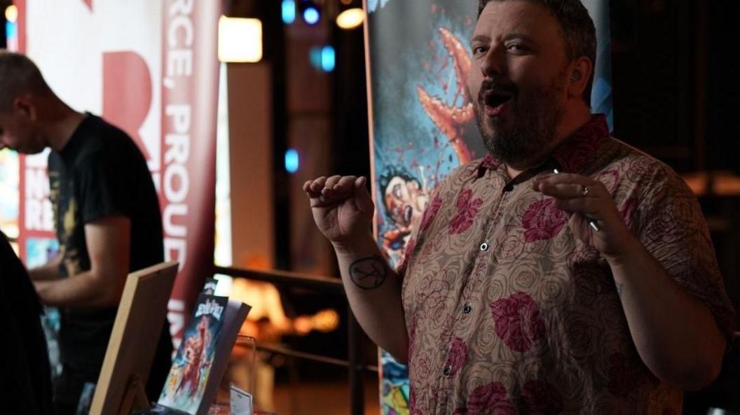 A man wearing a floral button-up shirt stands behind a stall with books on it. He is holding his hands up and has his mouth open as if in surprise.