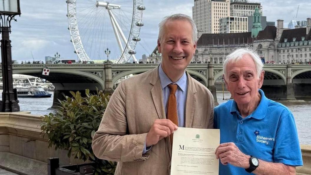 MP Gideon Amos wearing a pale blue collared shirt, orange tie and beige blazer. He is posing for a picture beside Brian Standring, who is wearing a blue branded Alzheimer's Society shirt and holding the document that shows his name was put forth for recognition. They are standing on a stone balcony outside the House of Commons, with the London Eye visible in the background.