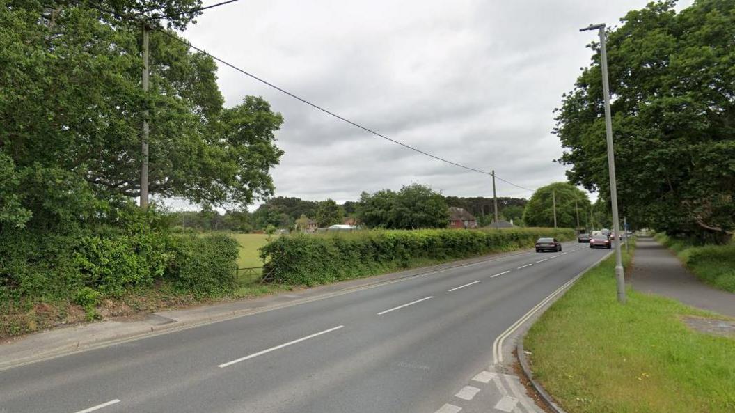 A Google Street View of an A road. A hedge runs alongside the left of the road, with a field beyond. On the right is a footpath.