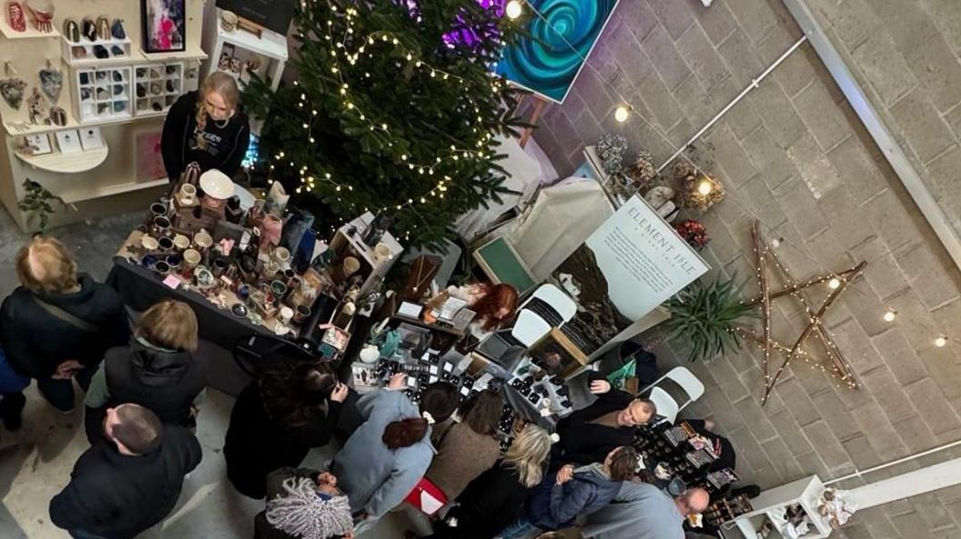 A Christmas fete with tables selling art, ceramics and jewellery. Traders sit behind the tables, people in winter outfits stand on the other side looking at the wares. A Christmas tree can be seen in the corner and a wooden star with twinkly lights is attached to a wall.