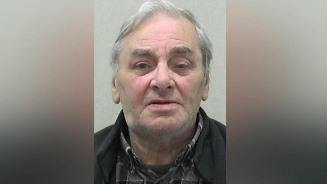 Mugshot of Gordon Hamilton. He has very faint grey stubble, one eye is more closed than the other and he has medium-length thin grey hair.