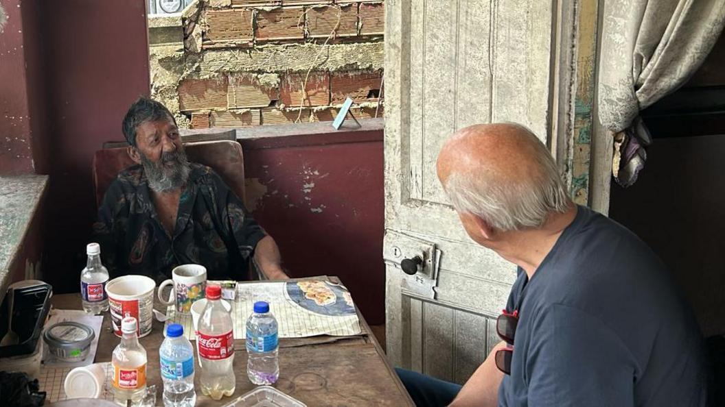 Nizam Hosein speaking to Ian McKay at Hosein's house in Trinidad. They both sit at a table filled with plastic bottles of water and coca cola. It is a porch with open windows on either side and is quite run down. Nizam wears an open necked dark shirt with paisley patterns and has a long bushy beard and short dark hair. Ian is seen from behind wearing a blue/grey t-shirt with red sunglasses clipped onto it.