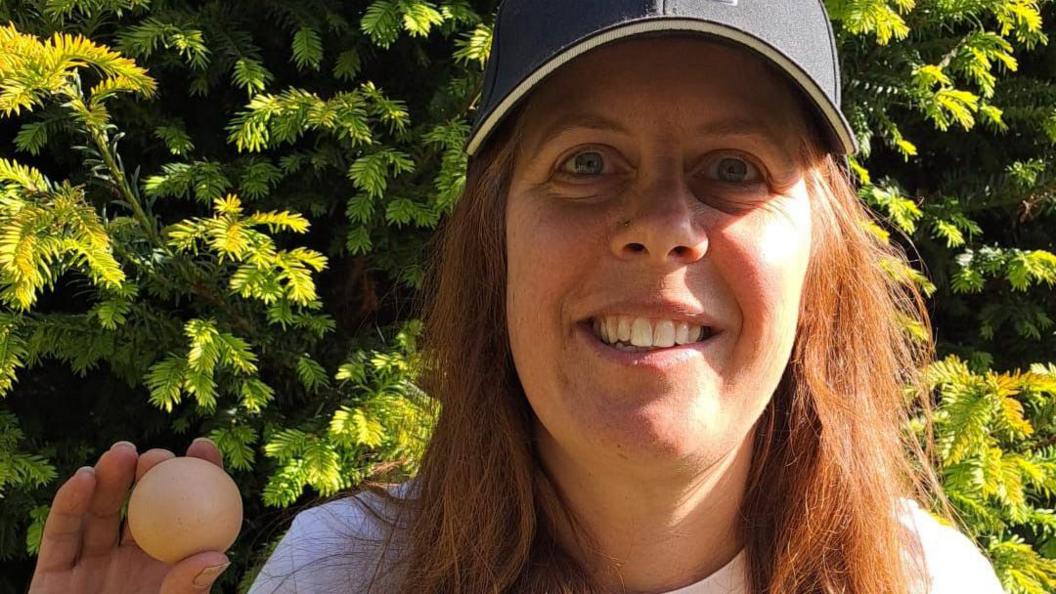 Jo Willis holds the egg in her hand. She is smiling at the camera as she stands outside in front of a large green bush. She wears a navy cap, black glasses, a white top and has long brown hair.