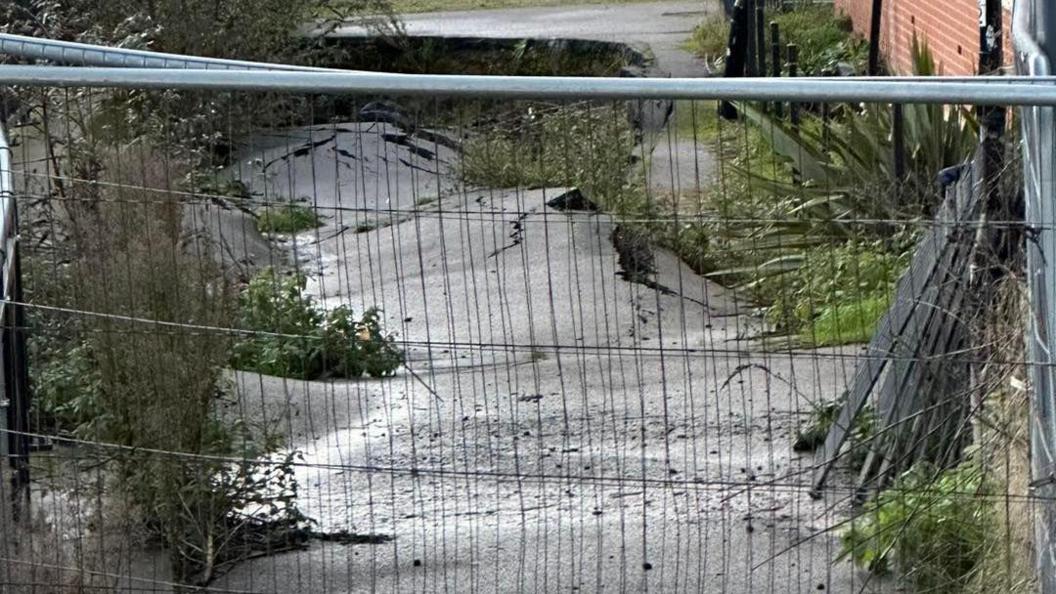 The footpath now buckled and cracked with vegetation growing out of the gaps in the tarmac, the path is fenced off from the public with Heras building fences about 2m (6ft) high.