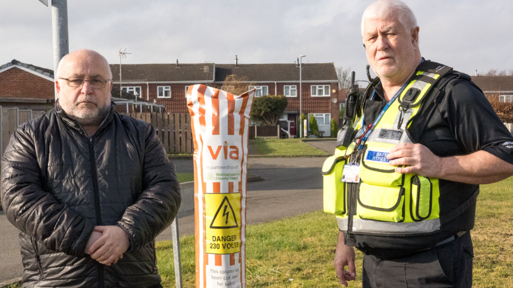 A man in a leather jacket and a police officer standing on either side of a CCTV pole with a protective sleave on it