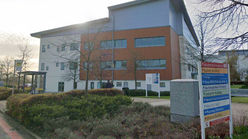 An exterior shot of Tameside Hospital. It is a modern, grey, three-storey building with landscaping in front and a directory of departments on a sign in the foreground.