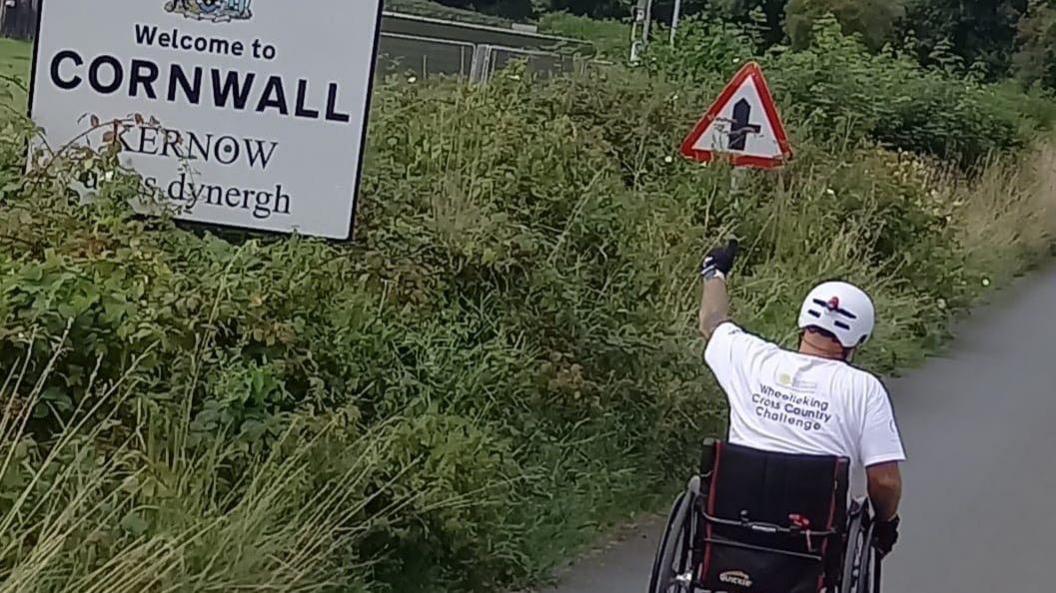 Ben Parker wearing a white t-shirt travelling away from the camera in his wheelchair. He is passing a sign that says "Welcome to Cornwall".