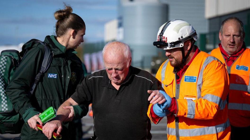 A paramedic and two search and rescue officers help a man who looks in distress. The female paramedic, dressed in green, and a male search and rescue officer, dressed in orange hi viz and a white hard hard, hold the man's arms as they help him walk. The man, who has grey hair and a black t-shirt, is looking down as though he is upset.