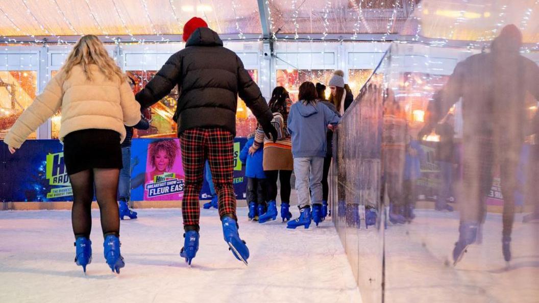 A couple skating on an ice rink as several children stand by the side of the rink behind them. There are fairy lights up above.