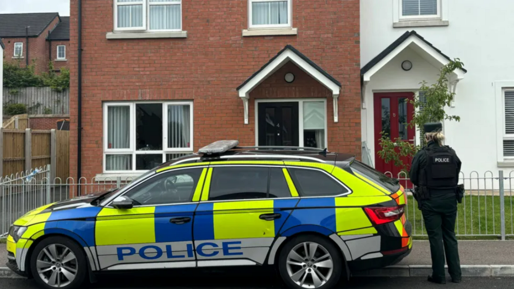 A police car is parked outside a red brick house with a female police officer standing to the right of the car