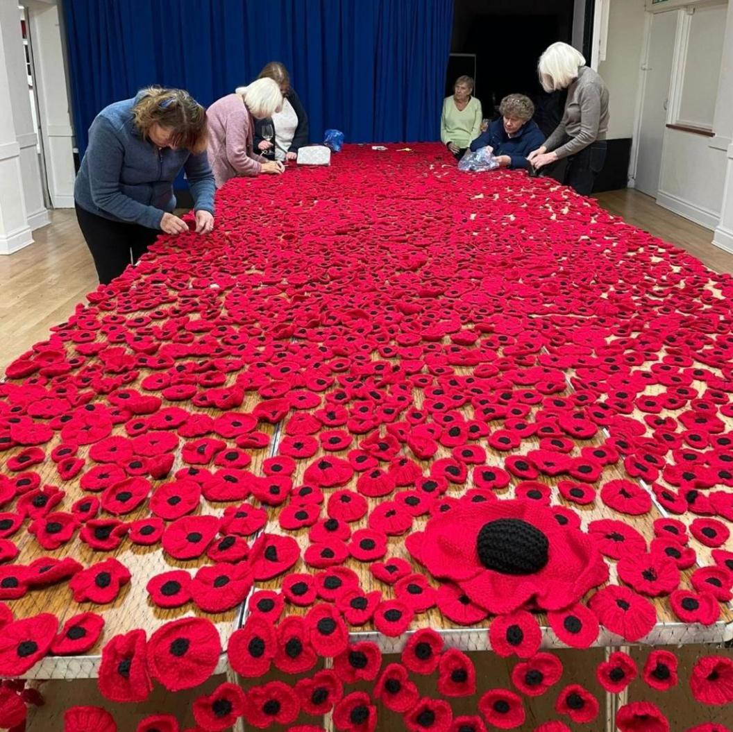 Five women put the finishing touches to a sea of red poppies laid out on a very long wooden table, while another woman watches them from the background