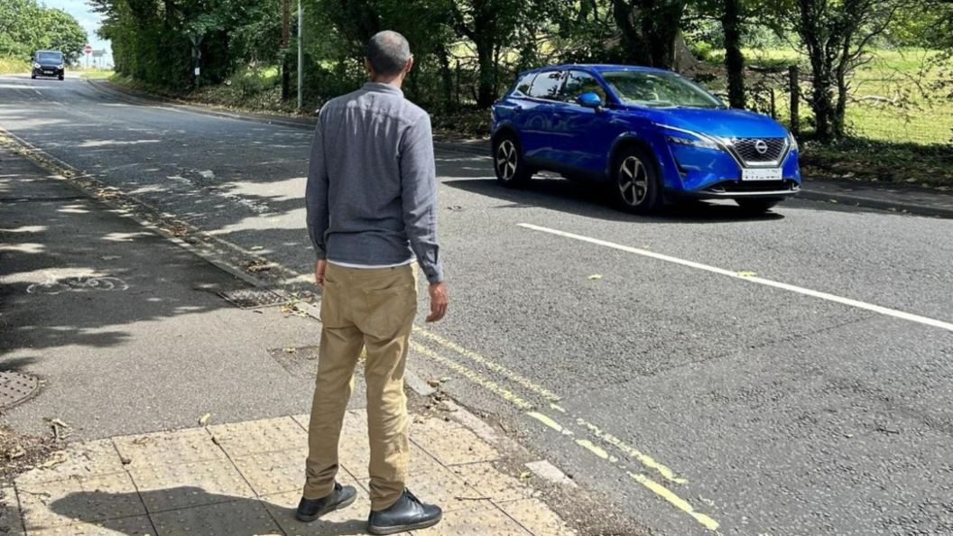 A man wear brown trousers and a blue shirt trying to cross a busy A road