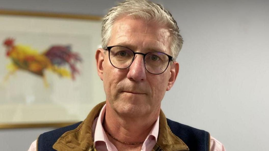 Mark Gorton. He is wearing black rimmed glasses, and a gilet over a pink patterned shirt. He has short grey hair and a serious look on his face.